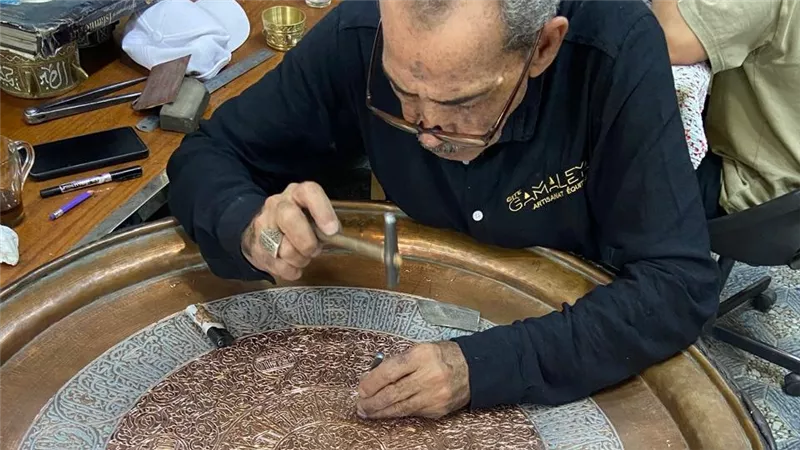 حرفي مصري ينقش الزخارف على صينية نحاسية باستخدام أدوات تقليدية، في حرفة تعتمد على الخبرة والدقة والعمل اليدوي. المصدر: الخبر المصري.